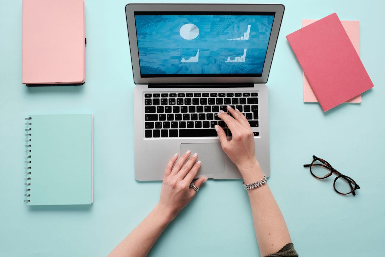 Hands typing on a laptop displaying data charts, with pink and teal office accessories.
